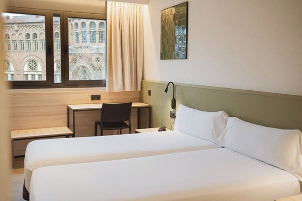 A hotel room with two beds, a desk, a chair, a lamp, and a window with a view of a building.