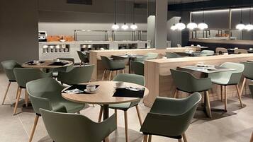 Een modern restaurant met ronde tafels en groene stoelen, een centraal gelegen bar en hanglampen.