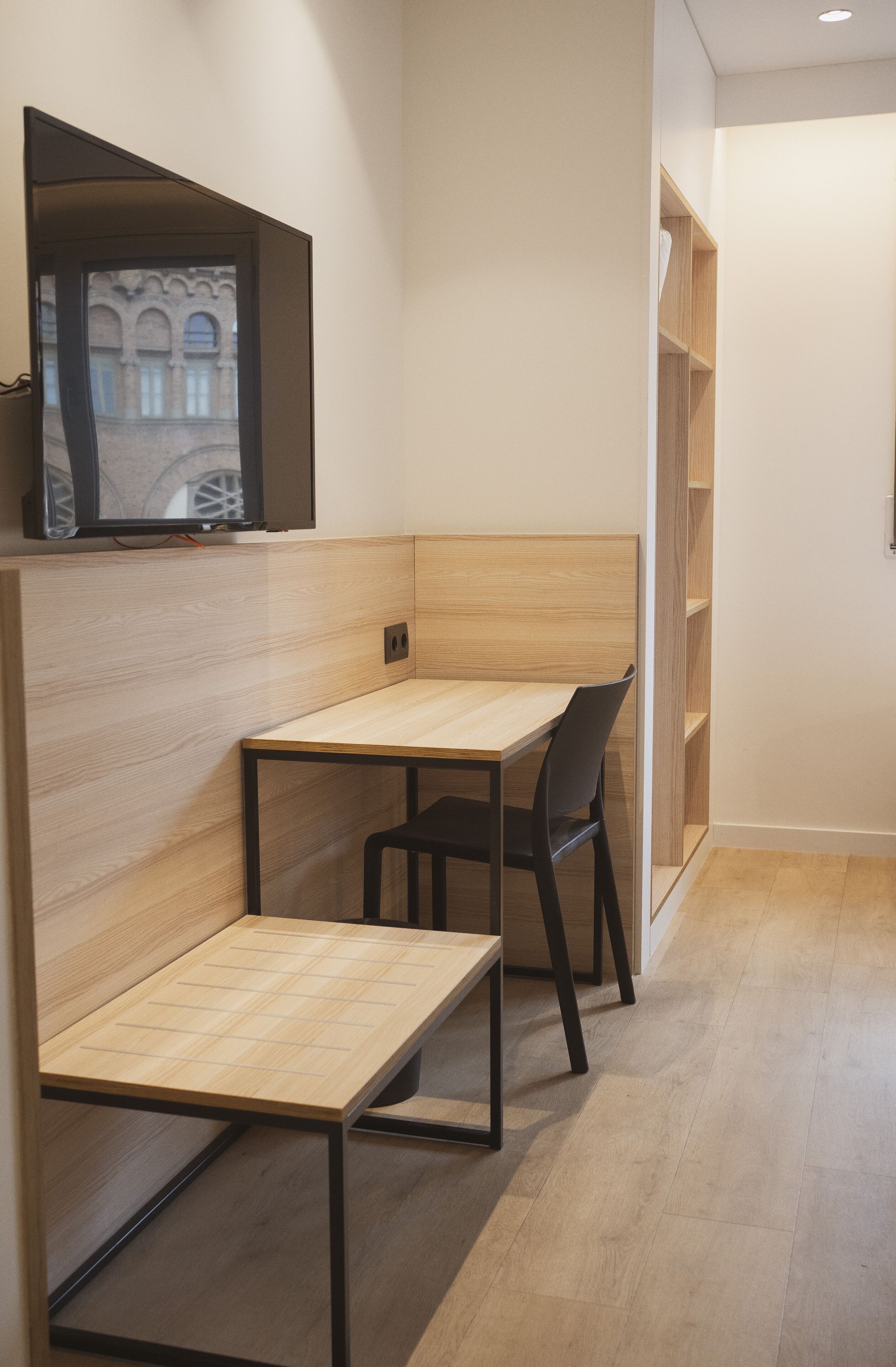 A modern room with a wooden desk and chair, a television mounted on the wall, and a bookshelf.