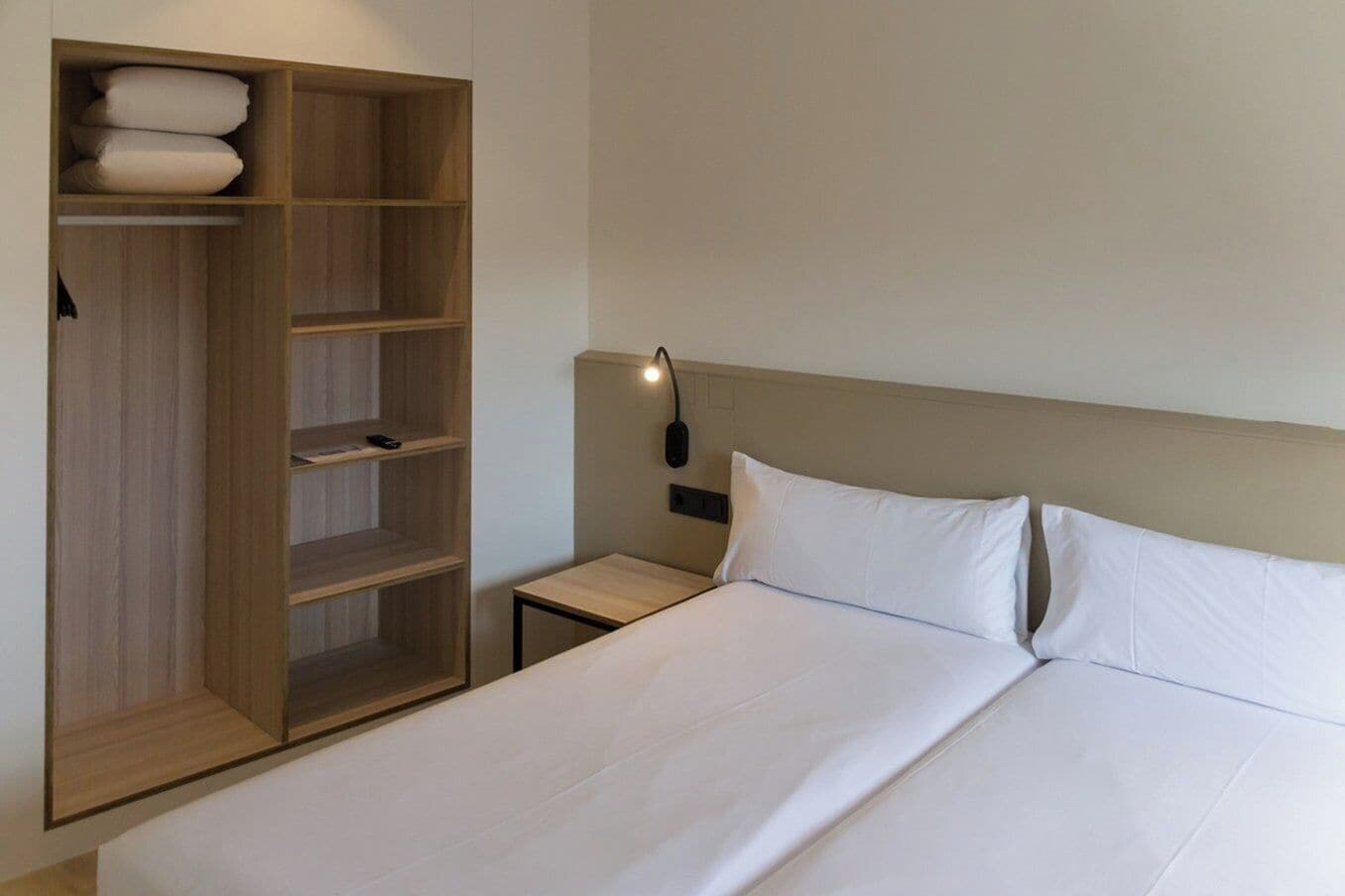 A hotel room with a bed, a wooden wardrobe, a bedside table, and a wall-mounted lamp.