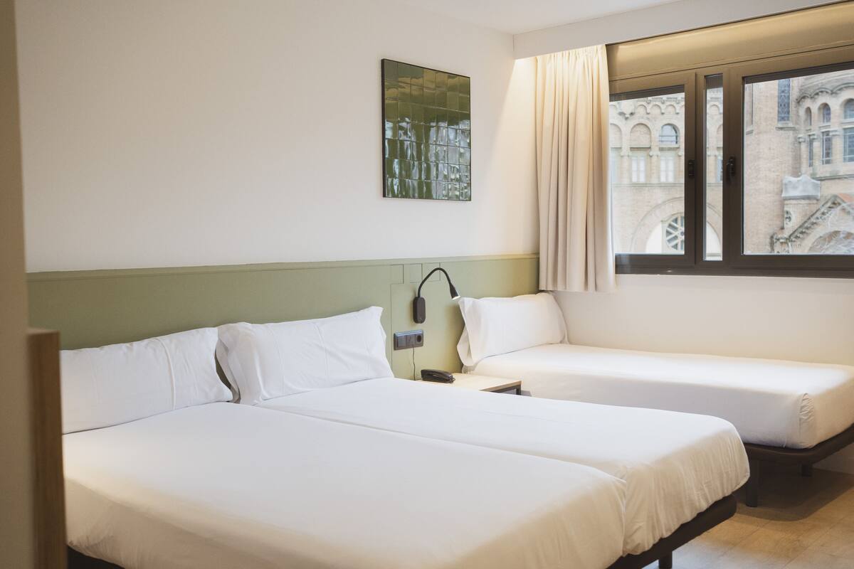 Habitación de hotel con dos camas, una mesita de noche con lámpara y ventana con vista a un edificio.