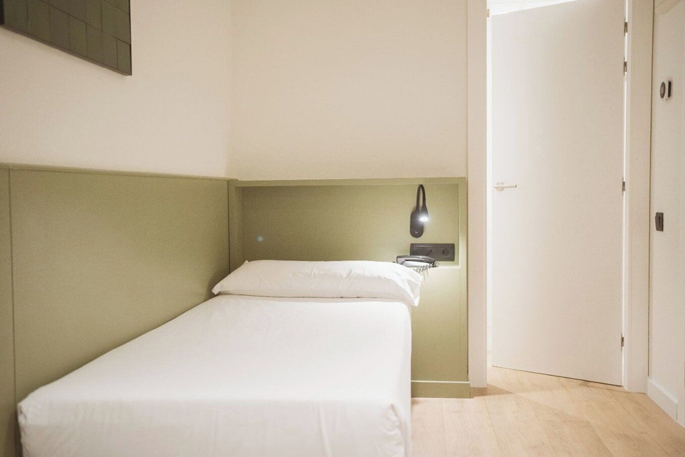 A small, single-bed room with a green headboard, a bedside table, and a wall-mounted lamp.
