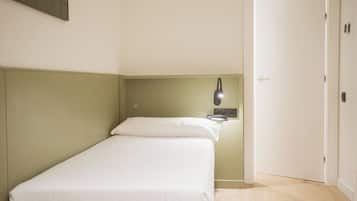 A small, single-bed room with a green headboard, a bedside table, and a wall-mounted lamp.