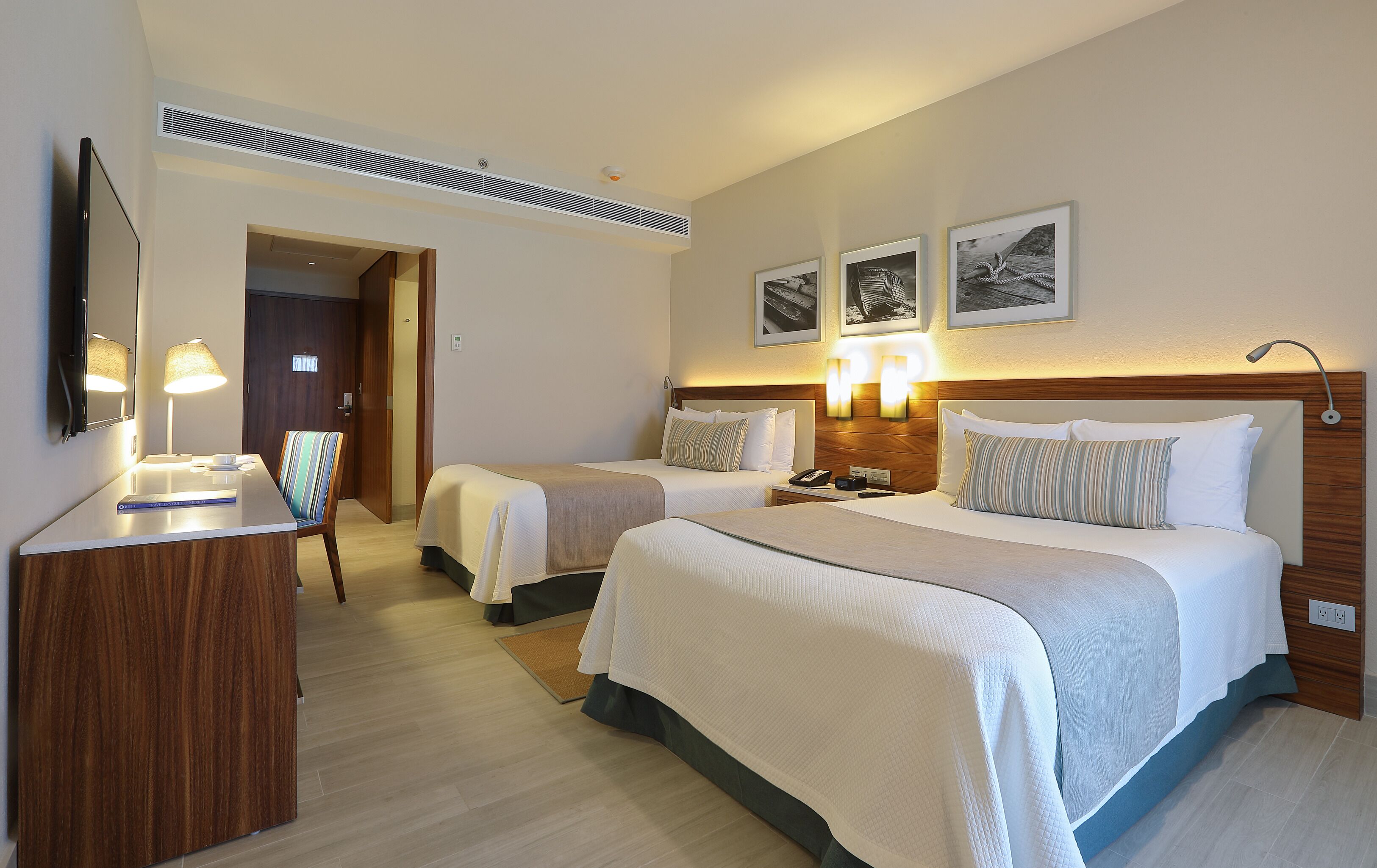 A hotel room with two beds, a desk, a chair, and a mirror. There are wall-mounted lights and a ceiling vent.
