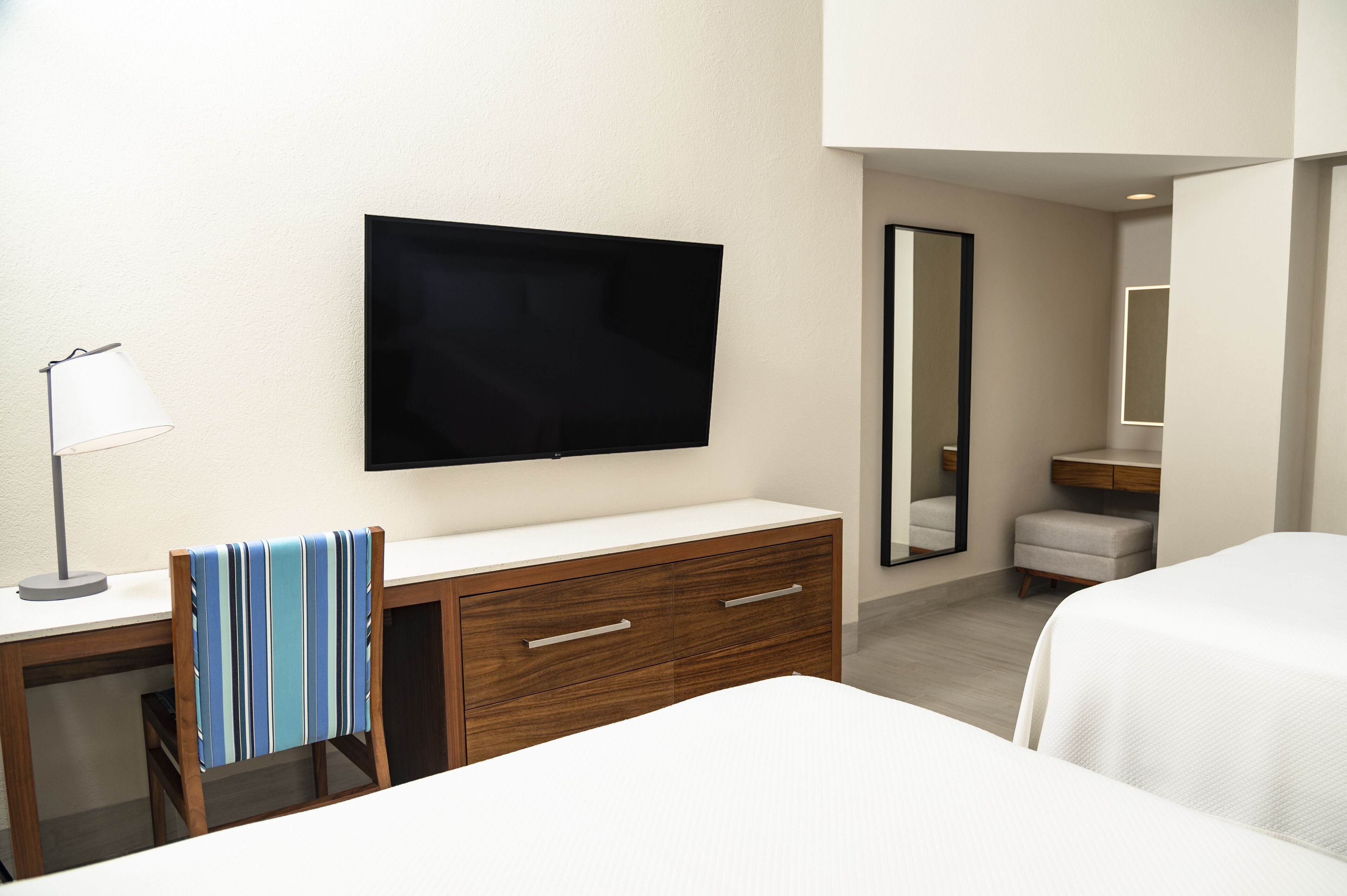 Een hotelkamer met een flatscreen televisie aan de muur, een houten bureau met lades, een gestreept fauteuil, een wit bed, een klein nachtkastje en een spiegel.
