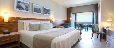 A hotel room with a large bed, a desk, a chair, and a balcony with a view of the sea.