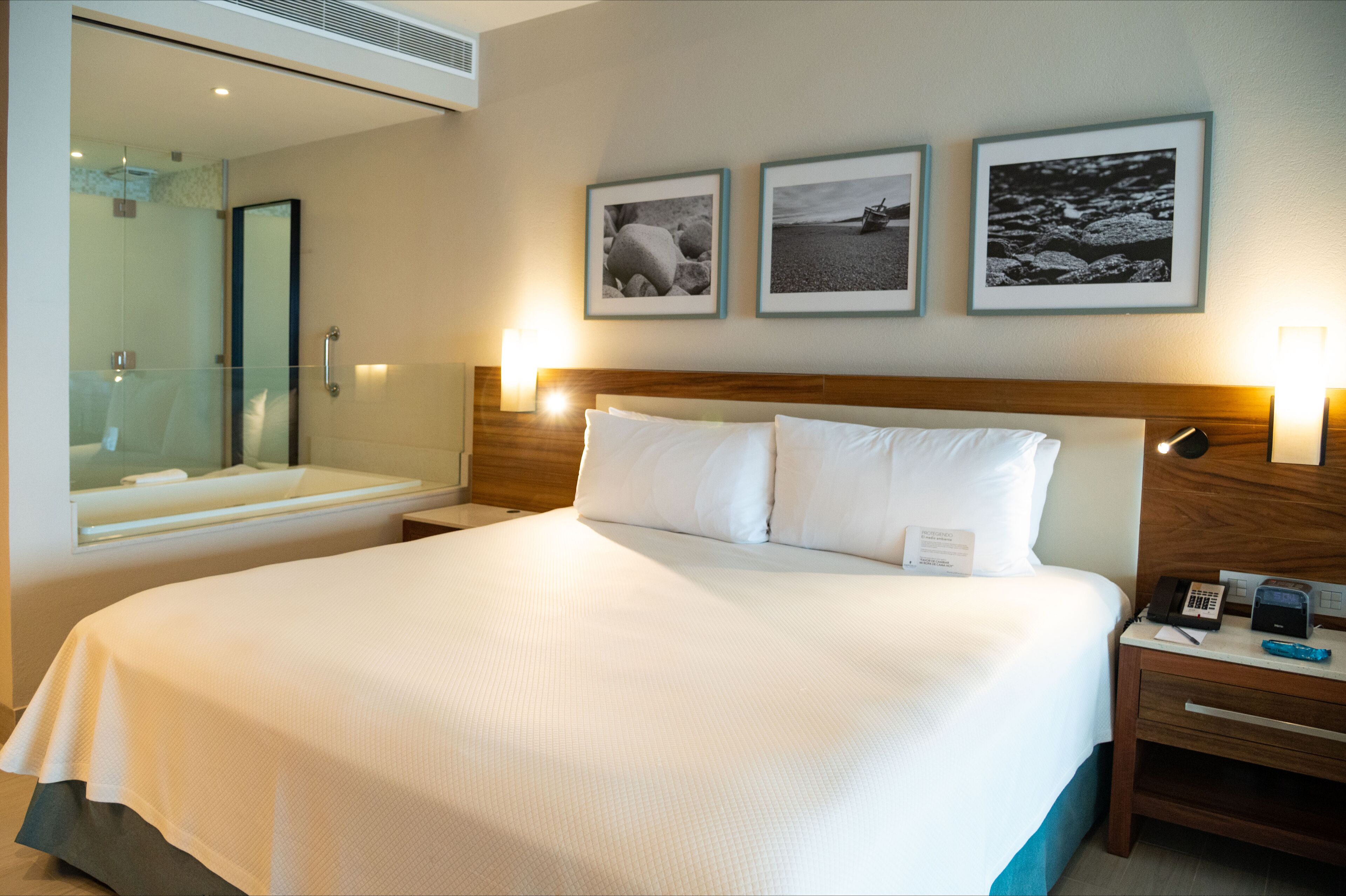 Habitación de hotel con una cama grande, cabecera de madera, mesita de noche con lámpara y espejo. Tres cuadros enmarcados adornan la pared sobre la cama.
