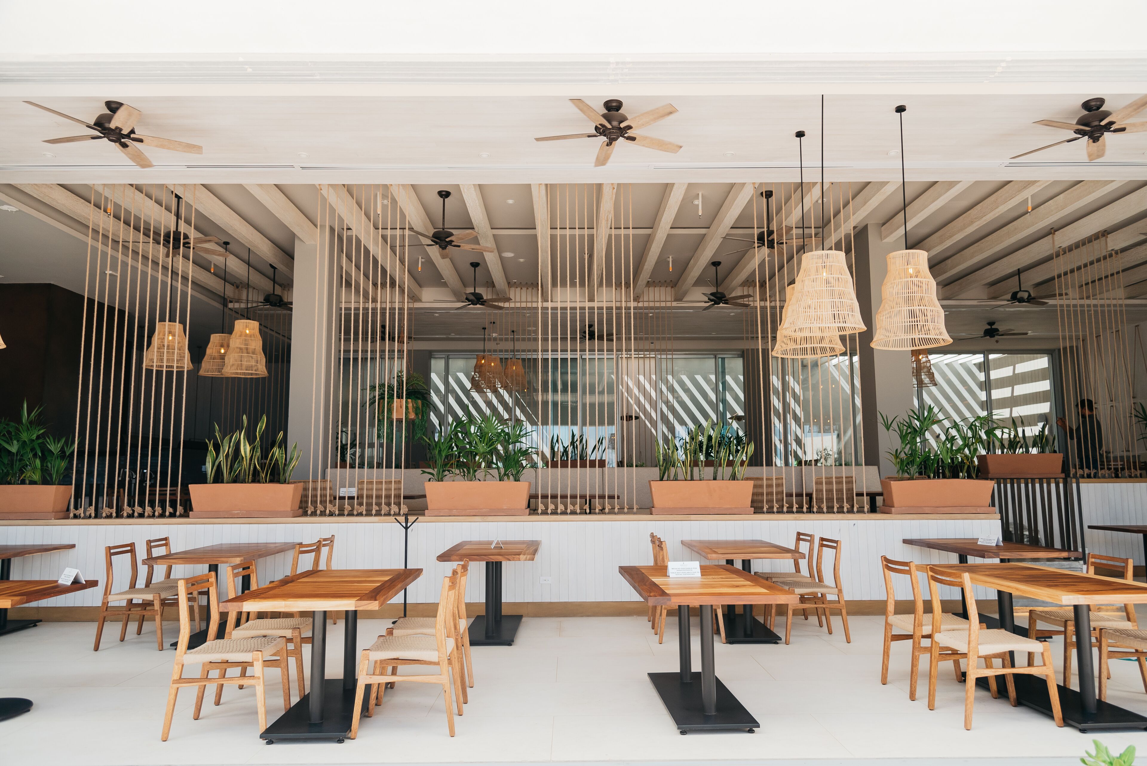 A modern cafe with wooden tables and chairs, ceiling fans, and hanging wicker lamps.