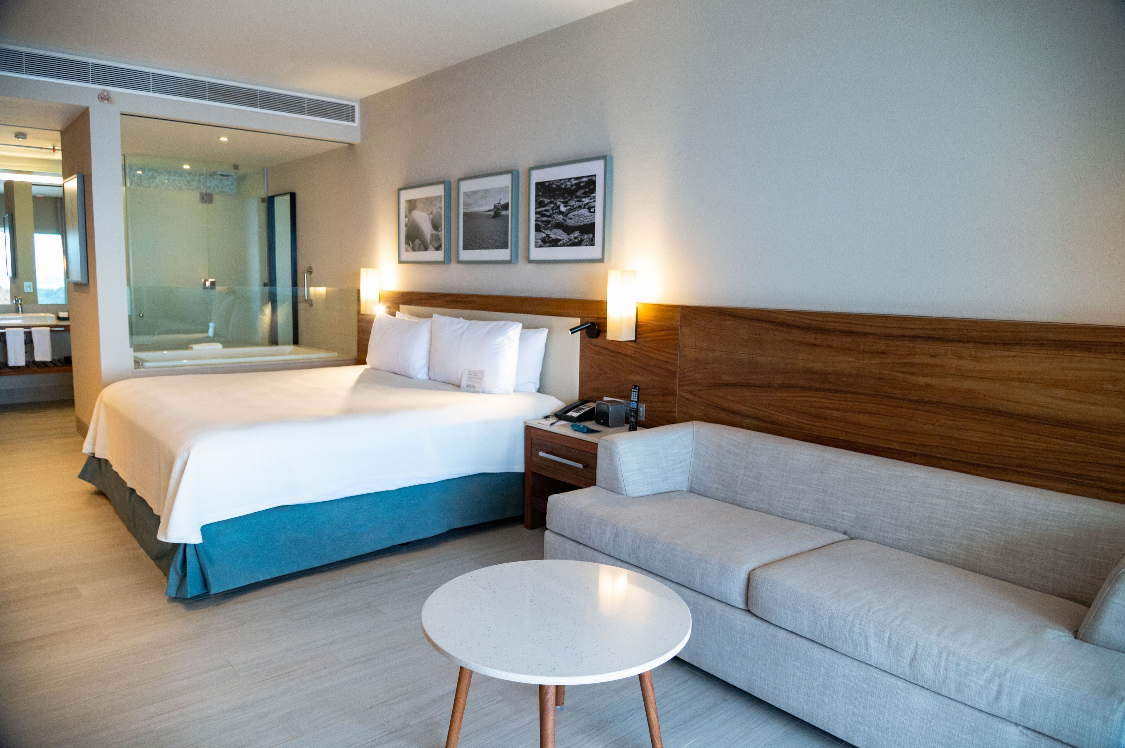 A hotel room with a bed, a sofa, a small table, a nightstand, a mirror, and a shower area.