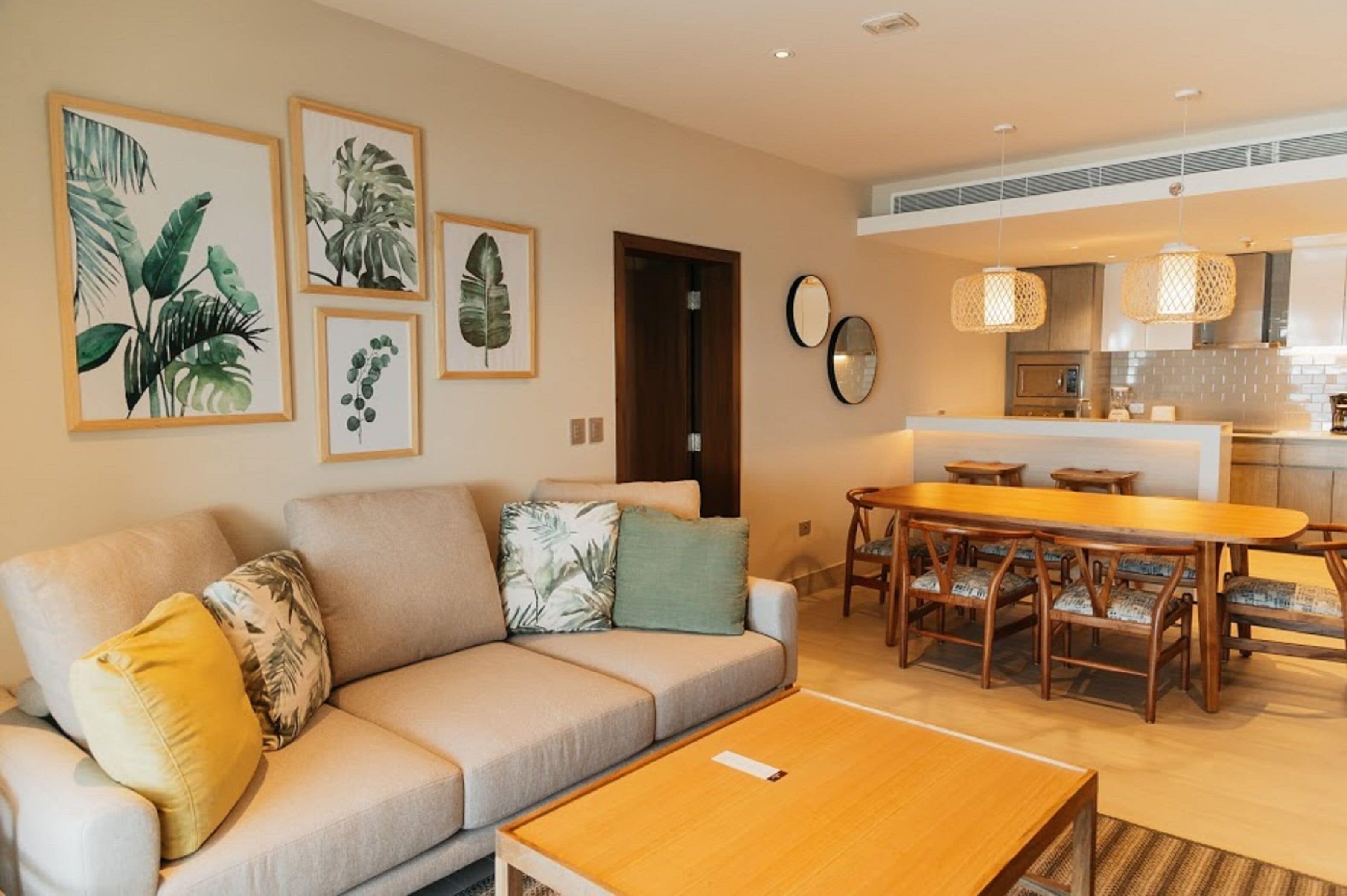 Een moderne woonkamer met een bank, salontafel en een eethoek. Op de muur hangen ingelijste botanische prints, er is een houten eettafel met stoelen, en er is een keuken zichtbaar door een open deur.