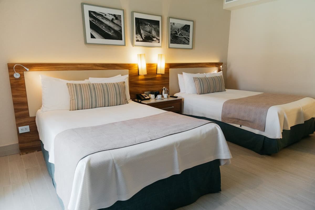 Habitación de hotel con dos camas, cada una con cabecero de madera, ropa de cama blanca y una mesita de noche con lámpara. Encima de las camas, hay tres cuadros enmarcados en la pared.