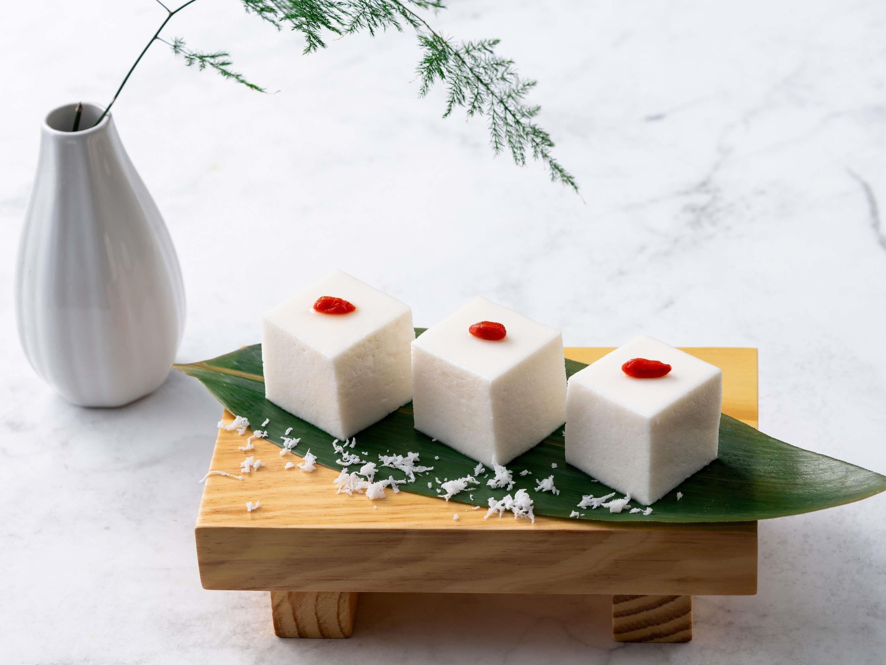 Three white square desserts on a wooden stand with a green leaf, a white vase, and a blurred background.