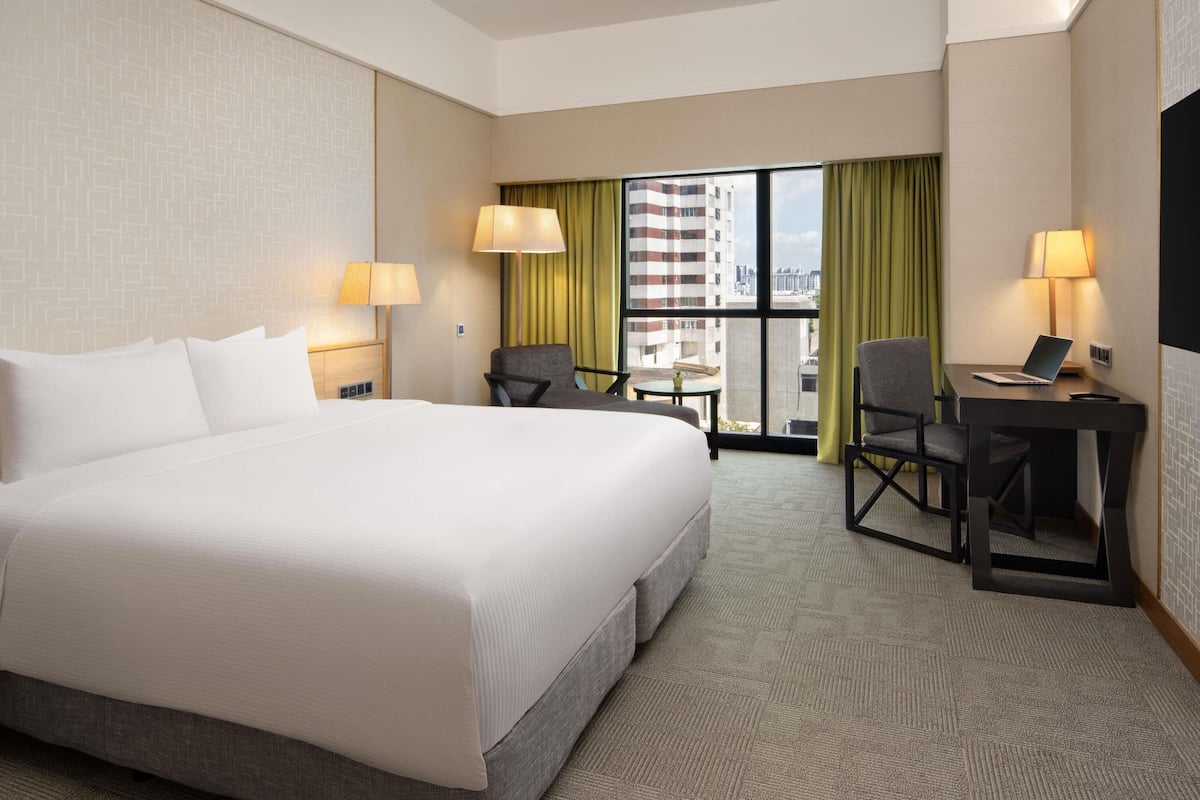 Habitación de hotel con una cama grande, dos sillas, un escritorio con una computadora portátil y vistas a un edificio a través de una ventana.