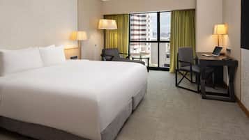 A hotel room with a large bed, two chairs, a desk with a laptop, and a view of a building through a window.
