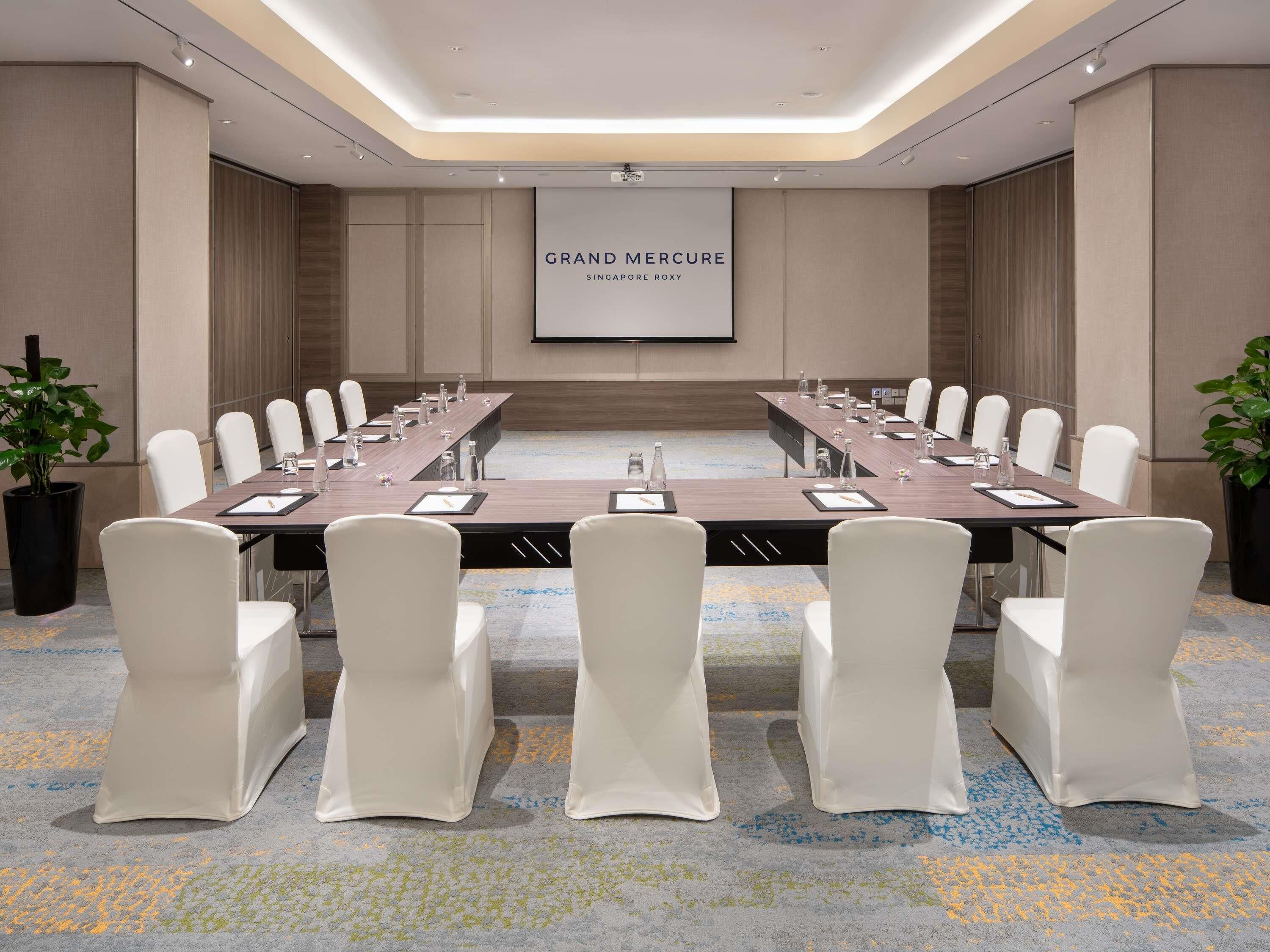 Ein Konferenzraum mit einem langen Tisch, weißen Stühlen und einer Projektorleinwand, die 'GRAND MERCURY SINGAPORE ROXY' zeigt.