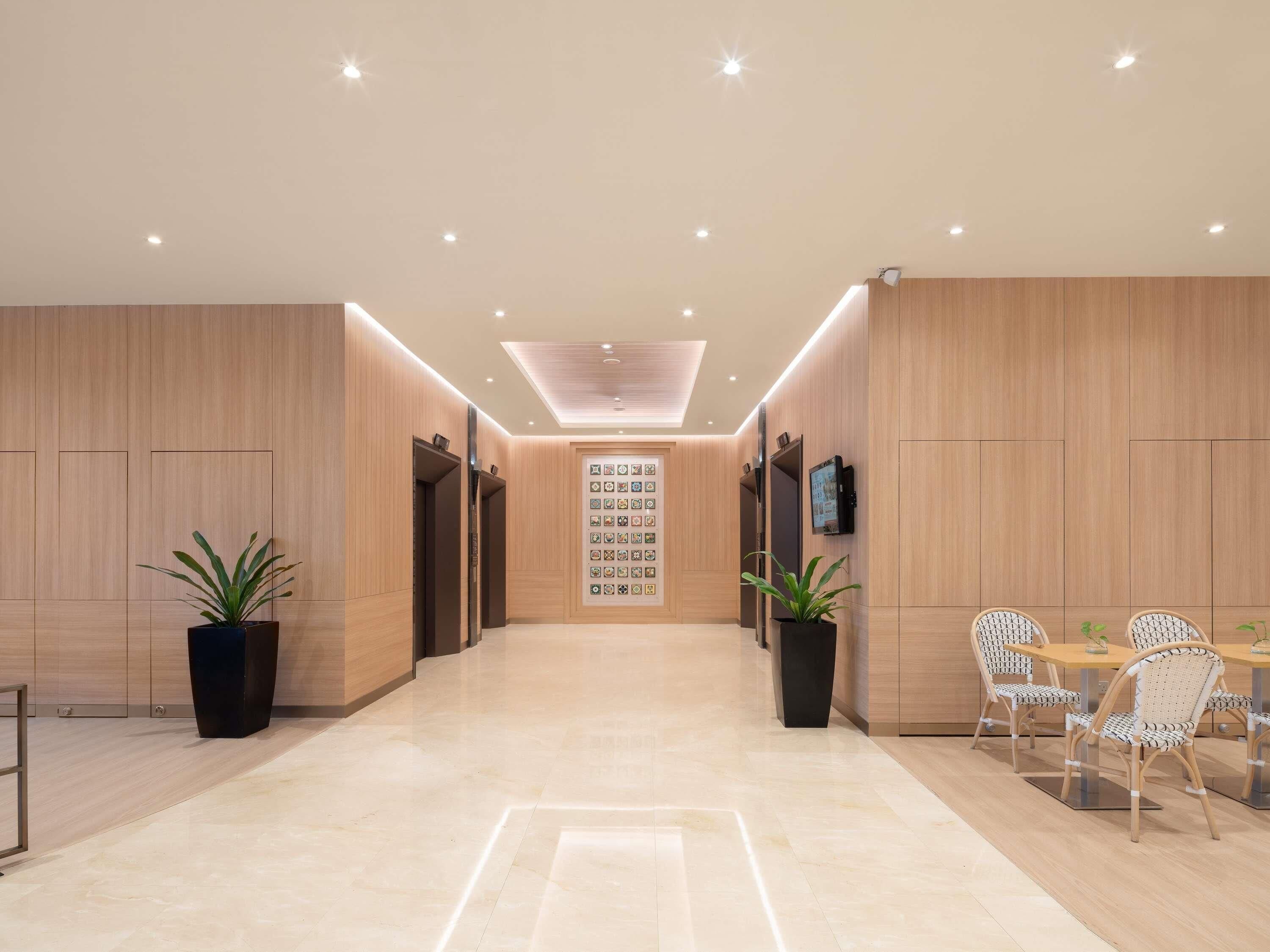 A modern hotel lobby with wooden paneling, a reception desk, two potted plants, a wall-mounted TV, and a dining area with wicker chairs.