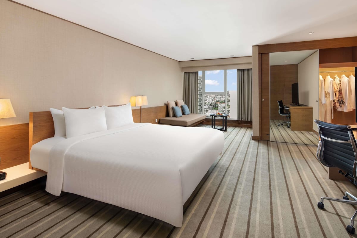 Habitación de hotel con una cama grande, un escritorio, una silla, un sofá y vistas de la ciudad a través de amplias ventanas.