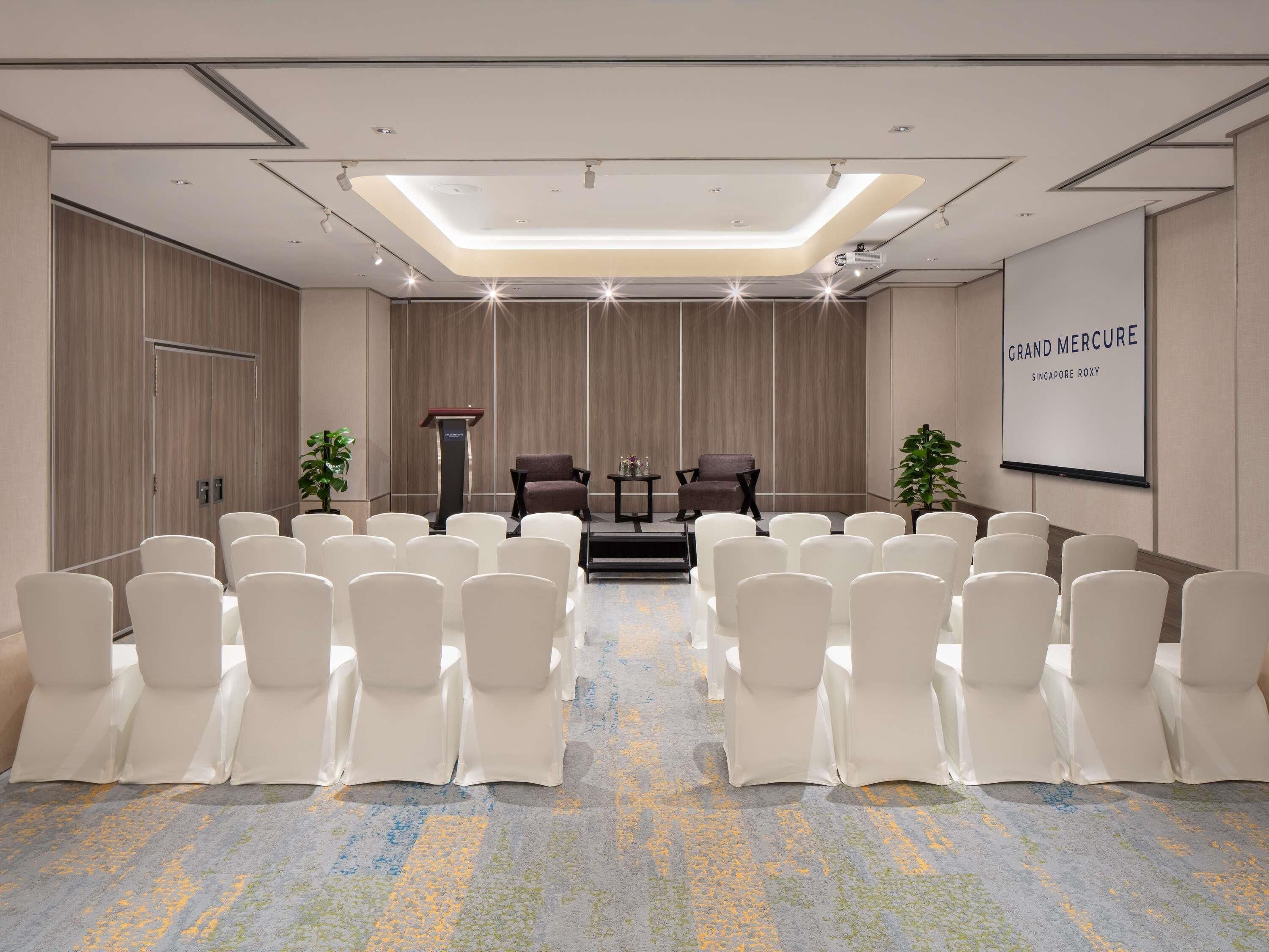 A conference room with rows of white chairs, a podium, two sofas, and a screen displaying 'GRAND MERCURE SINGAPORE ROXY'.