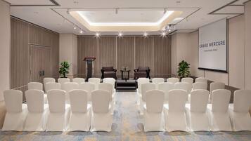 A conference room with rows of white chairs, a podium, two sofas, and a screen displaying 'GRAND MERCURE SINGAPORE ROXY'.
