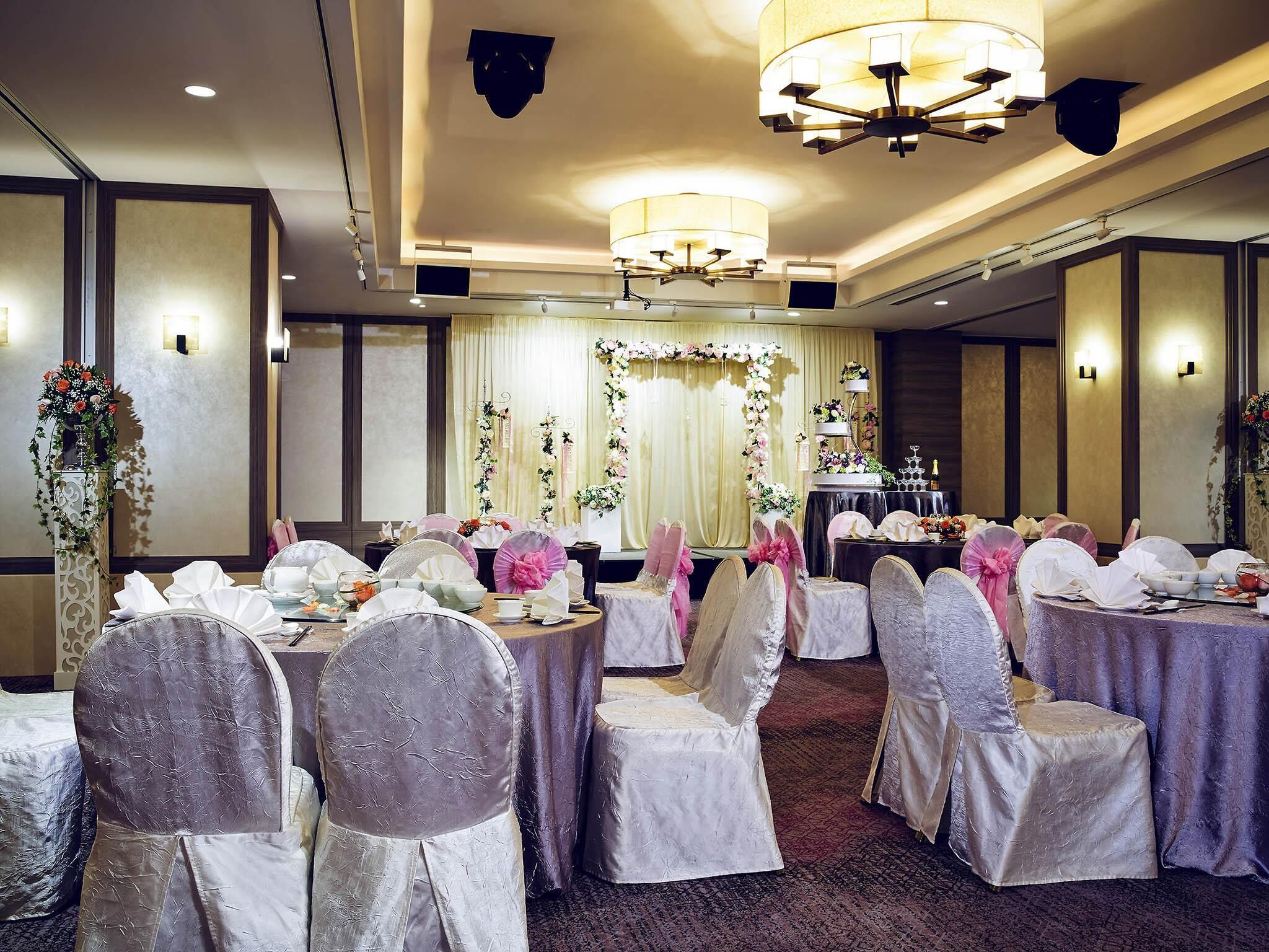 A banquet hall with round tables, chairs with white and pink decorations, and a stage with floral arrangements.