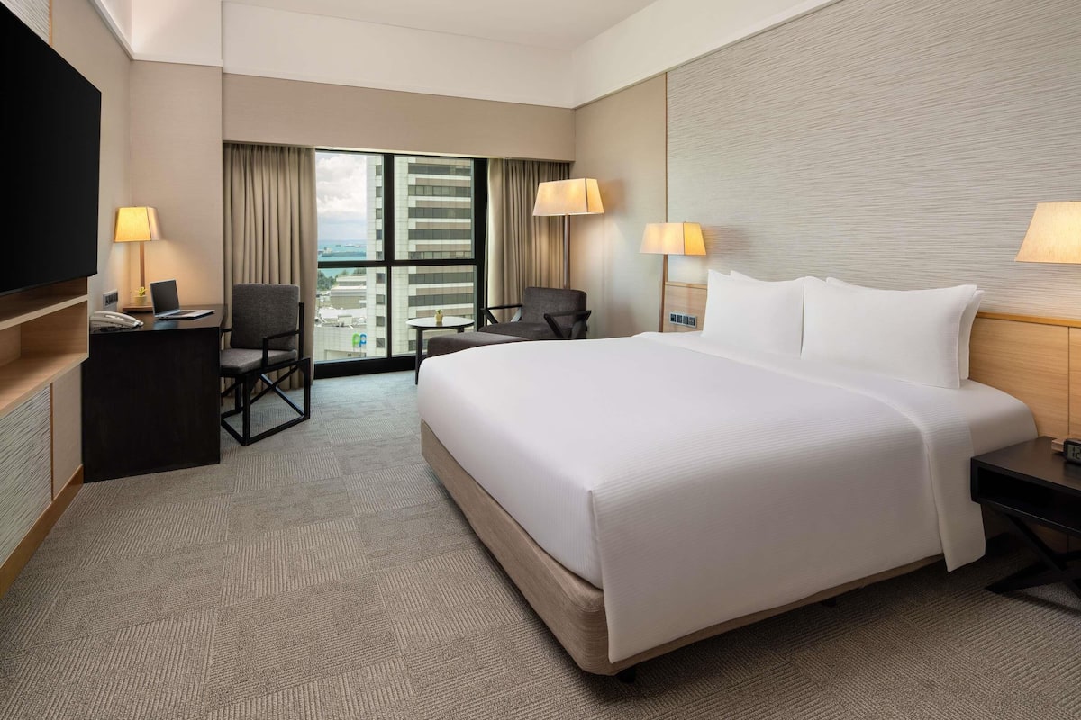 Habitación de hotel con una cama grande, un escritorio con lámpara, una silla, un televisor y vistas de la ciudad a través de amplios ventanales.
