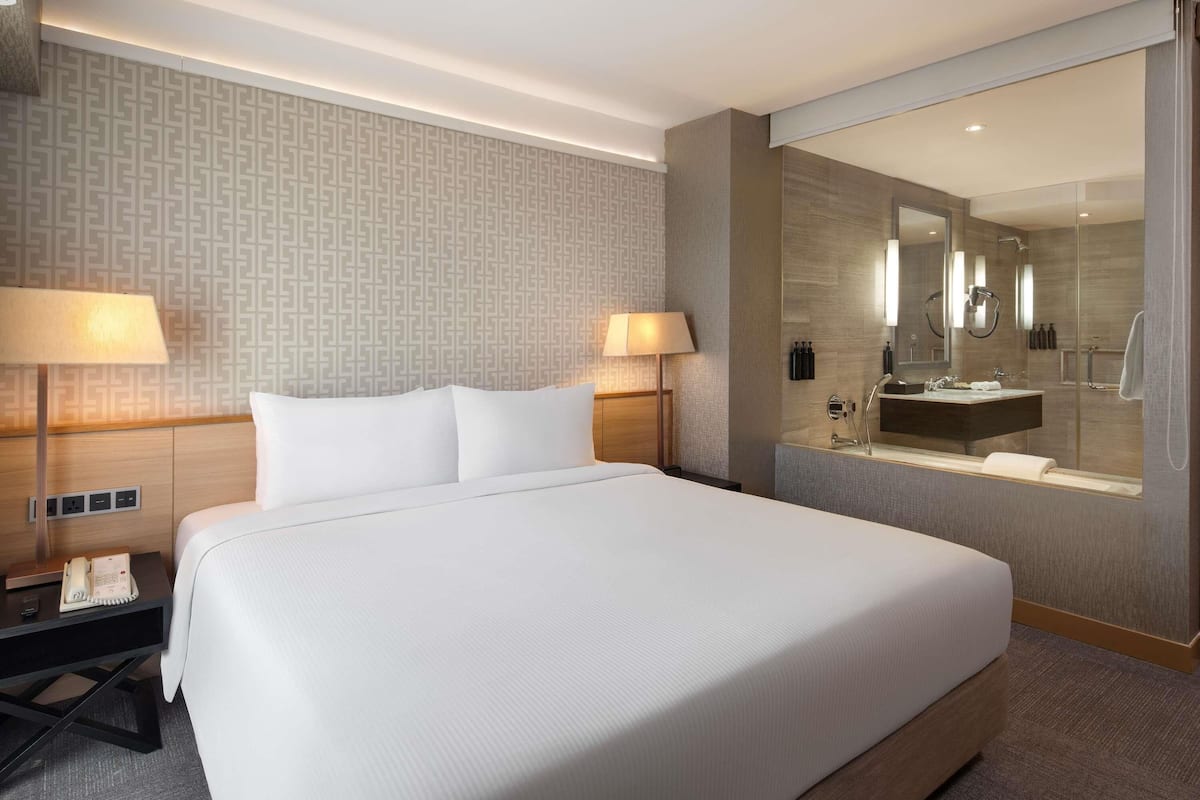 Habitación de hotel con una cama grande, lámparas de noche, una mesita de noche con teléfono, y un baño con espejo, lavabo y ducha.