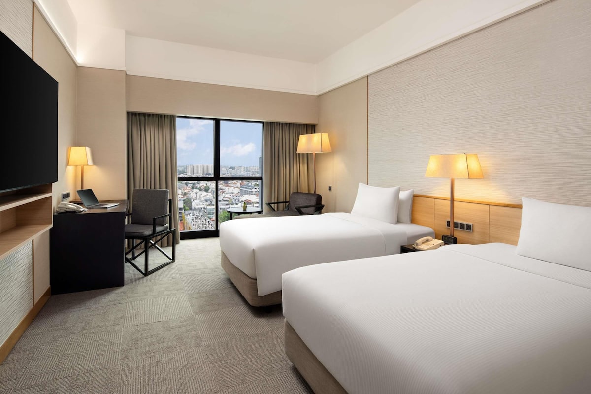 Habitación de hotel con dos camas, un escritorio con una computadora portátil, una silla, un televisor y vista a la ciudad.