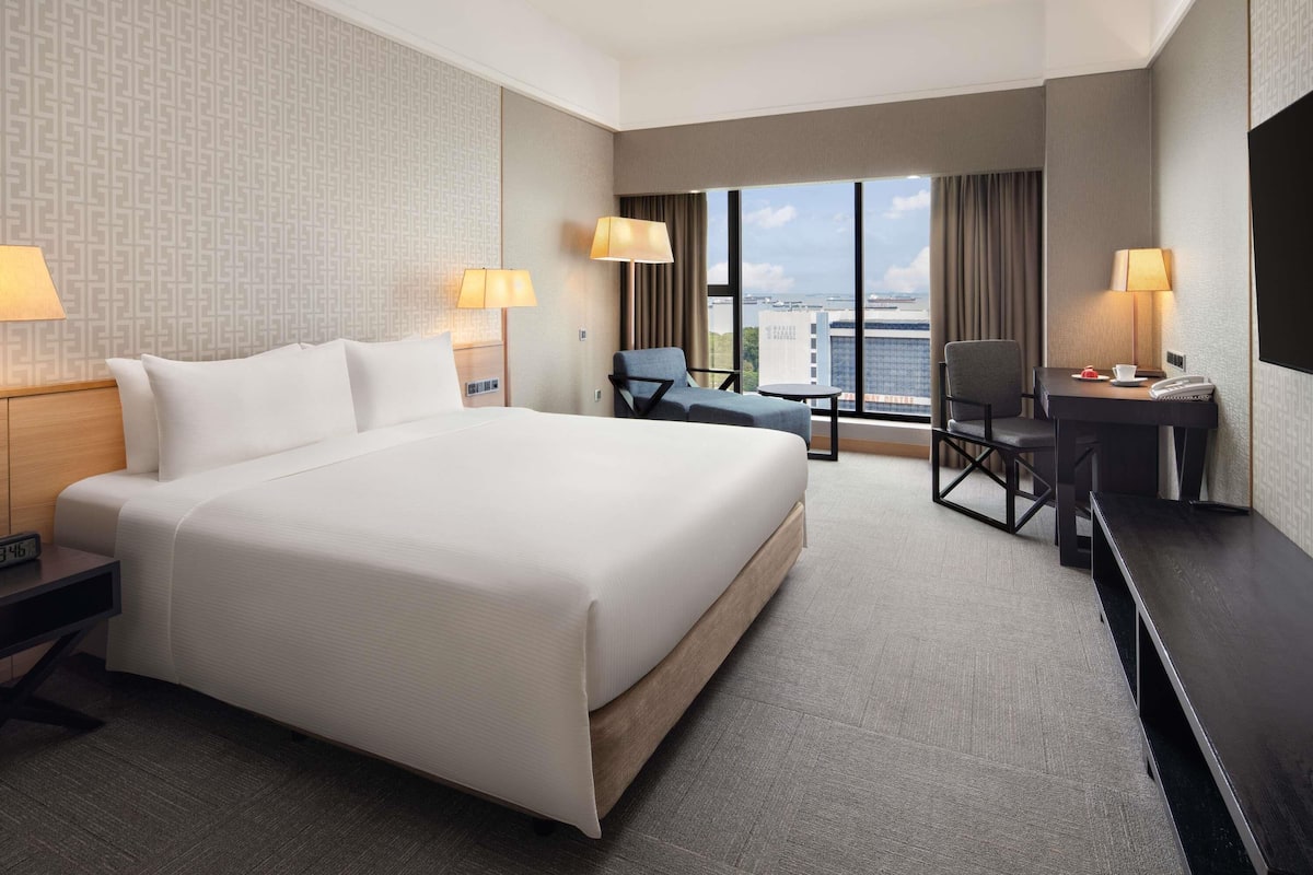 Habitación de hotel con una cama grande, dos mesitas de noche, un escritorio con silla, una mesita redonda y un ventanal con vistas a la ciudad.