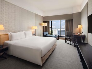 Quarto de hotel com uma cama grande, duas mesas de cabeceira, uma escrivaninha com cadeira, uma mesinha redonda e uma janela ampla com vista para a cidade.