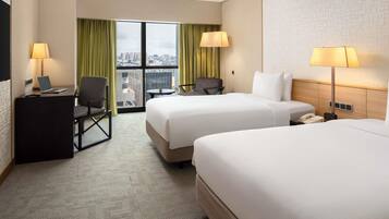 Ein Hotelzimmer mit zwei Betten, einem Schreibtisch mit einem Laptop, einem Sessel, einem kleinen Tisch und Blick auf die Stadt.