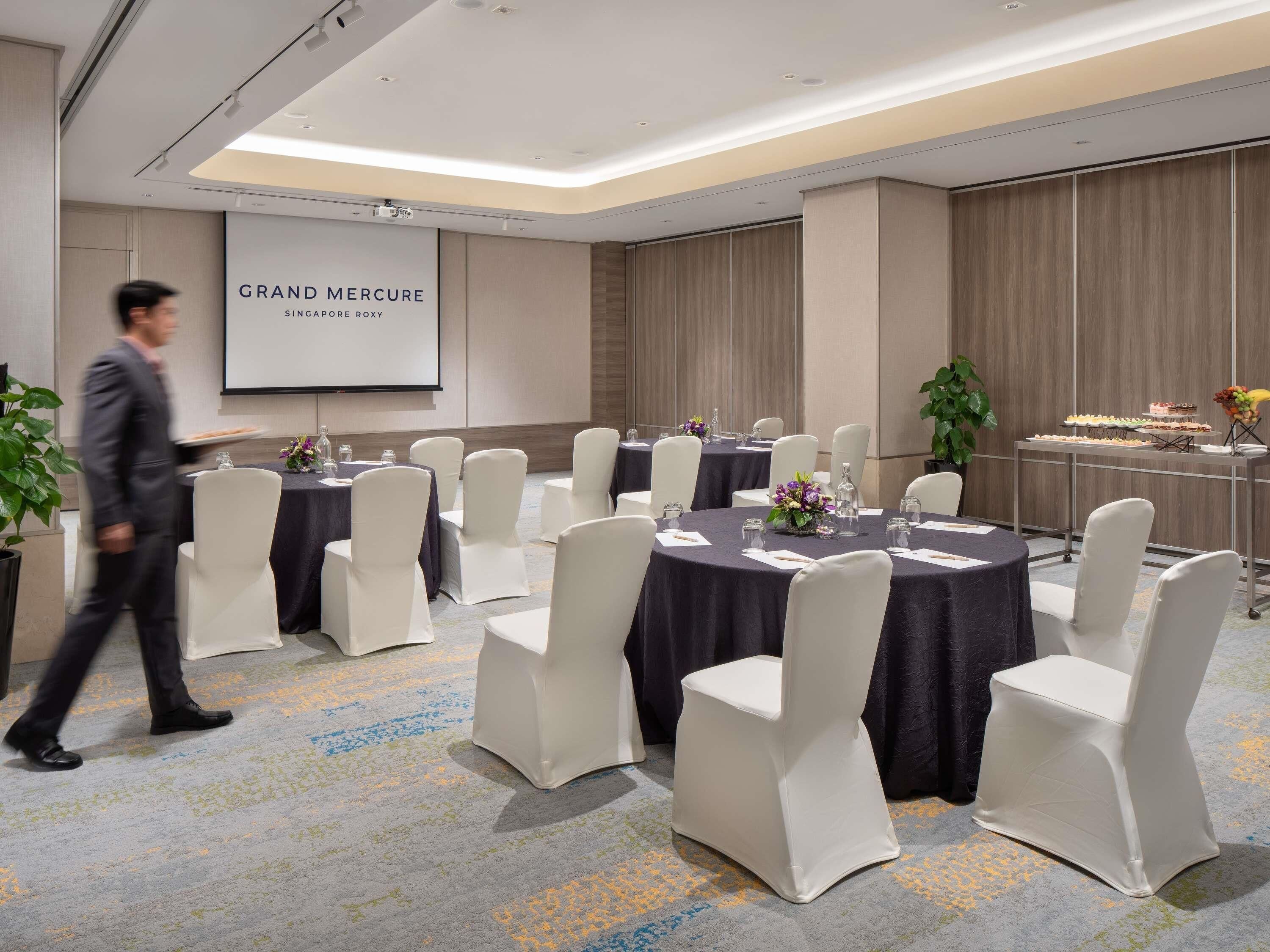 Un salón de conferencias con mesas redondas, sillas blancas, una pantalla de proyector y una mesa de buffet.