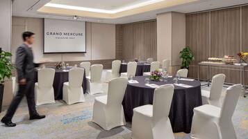 Un salón de conferencias con mesas redondas, sillas blancas, una pantalla de proyector y una mesa de buffet.