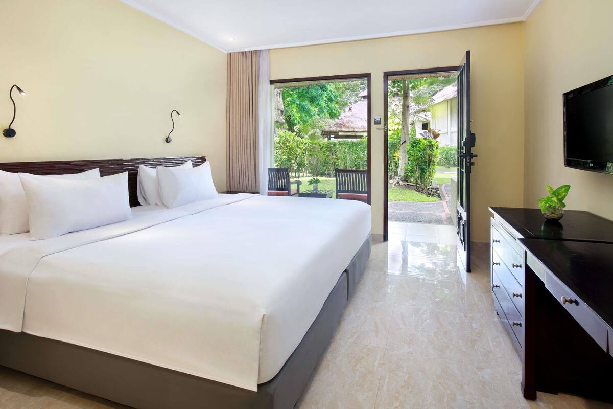 Ein Hotelzimmer mit einem großen Bett, einem Fernseher, einem Schreibtisch mit Schubladen und Blick auf einen Garten durch eine geöffnete Tür.