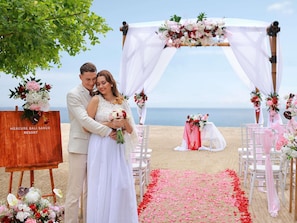 A wedding ceremony on a beach with a wooden easel labeled 'Mercure Bali Sanur Resort' and floral decorations.