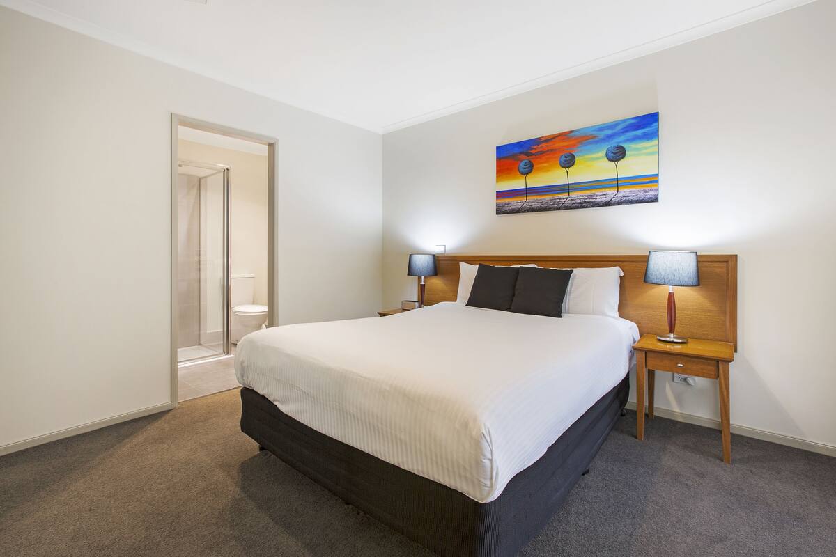 A bedroom with a large bed, two bedside tables with lamps, a painting on the wall, and a door leading to a bathroom.
