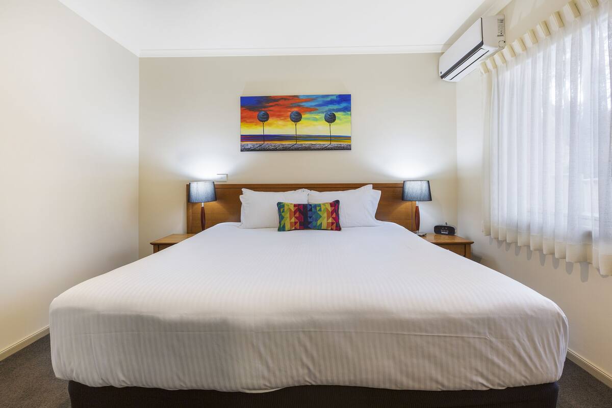 A hotel room with a large bed, two bedside lamps, a painting on the wall, and an air conditioning unit.