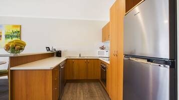 A compact kitchen with wooden cabinets, a stainless steel refrigerator, a microwave, and a bowl of fruit on the counter.
