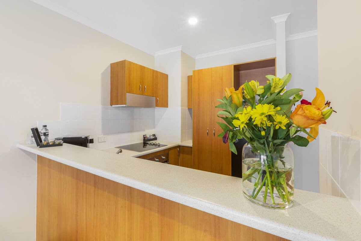 A modern kitchen with wooden cabinets, a white countertop, and a vase of flowers on the counter.