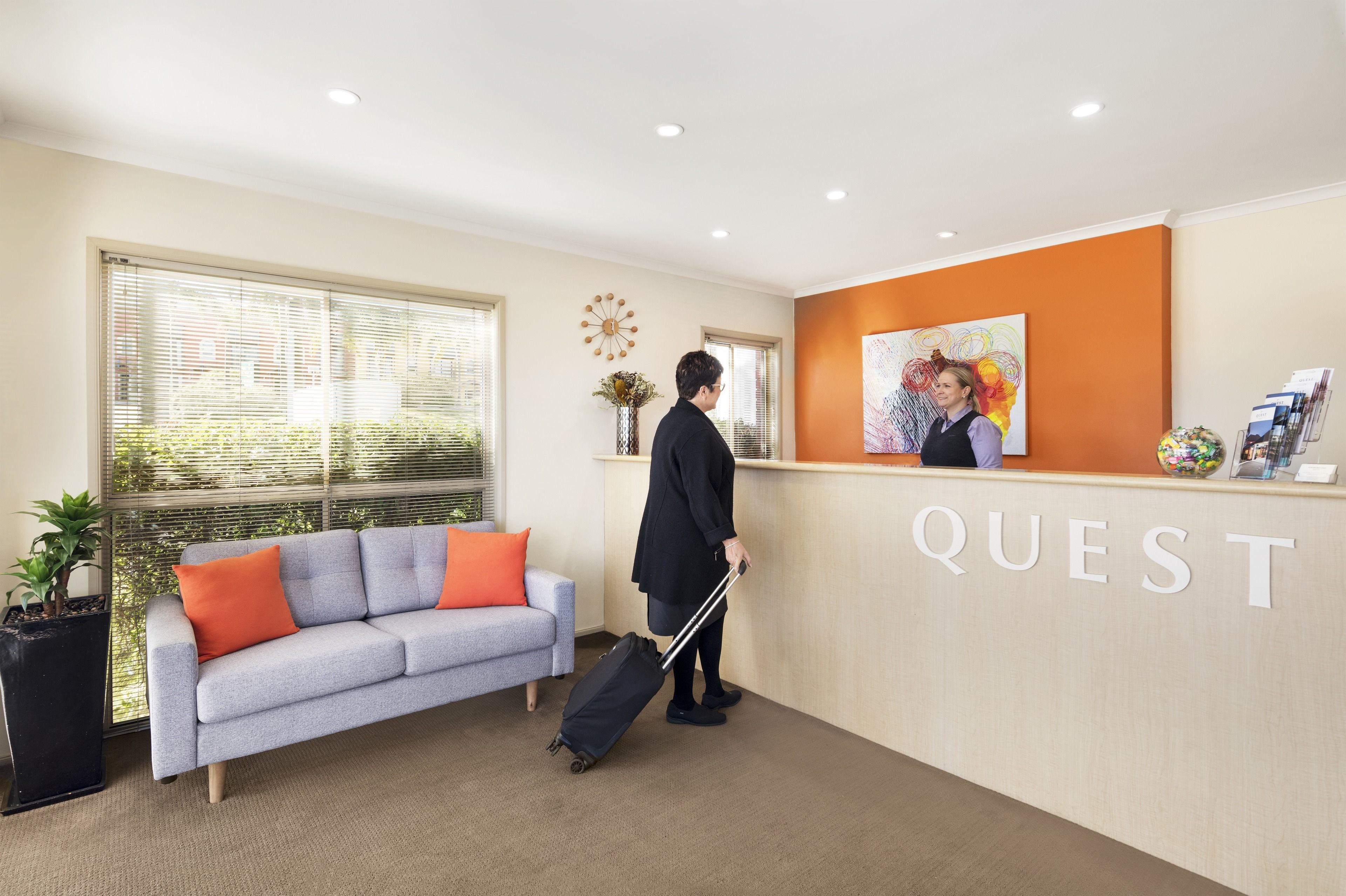 A reception area with a grey sofa, orange cushions, a black suitcase, and a 'QUEST' sign on the wall.