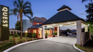 Entrada de un hotel con un camino cubierto, palmeras y un letrero que dice 'QUEST'.