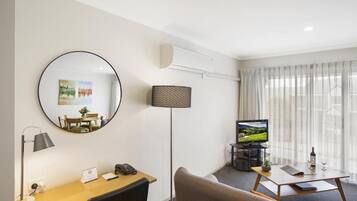 Ein modernes Hotelzimmer mit einem runden Spiegel, einem Schreibtisch mit Telefon, einer Lampe, einem Fernseher auf einem Podest und einem Sofa.