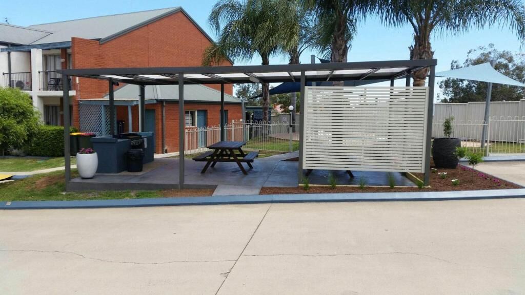 Un espacio al aire libre techado con mesa de picnic, un cubo de basura y una jardinera con flores.