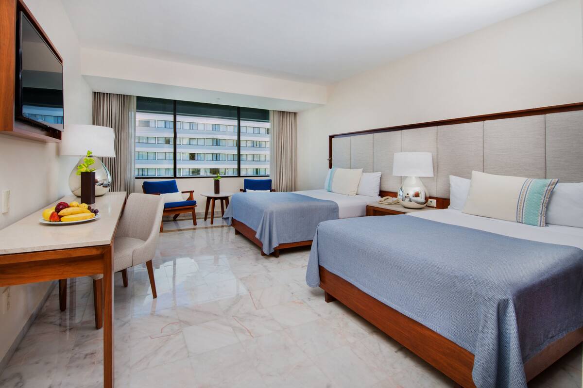 A hotel room with two beds, a desk, a chair, a TV, and a window with a view of a building.