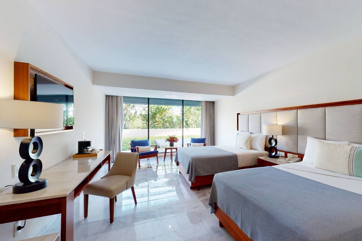 A hotel room with two beds, a desk with a lamp, a chair, a TV, and a large window offering a view of greenery.