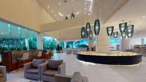 A spacious lobby with wicker furniture, a central water feature, and large windows offering a city view. - Park Royal Beach Cancun - All Inclusive (Cancun)