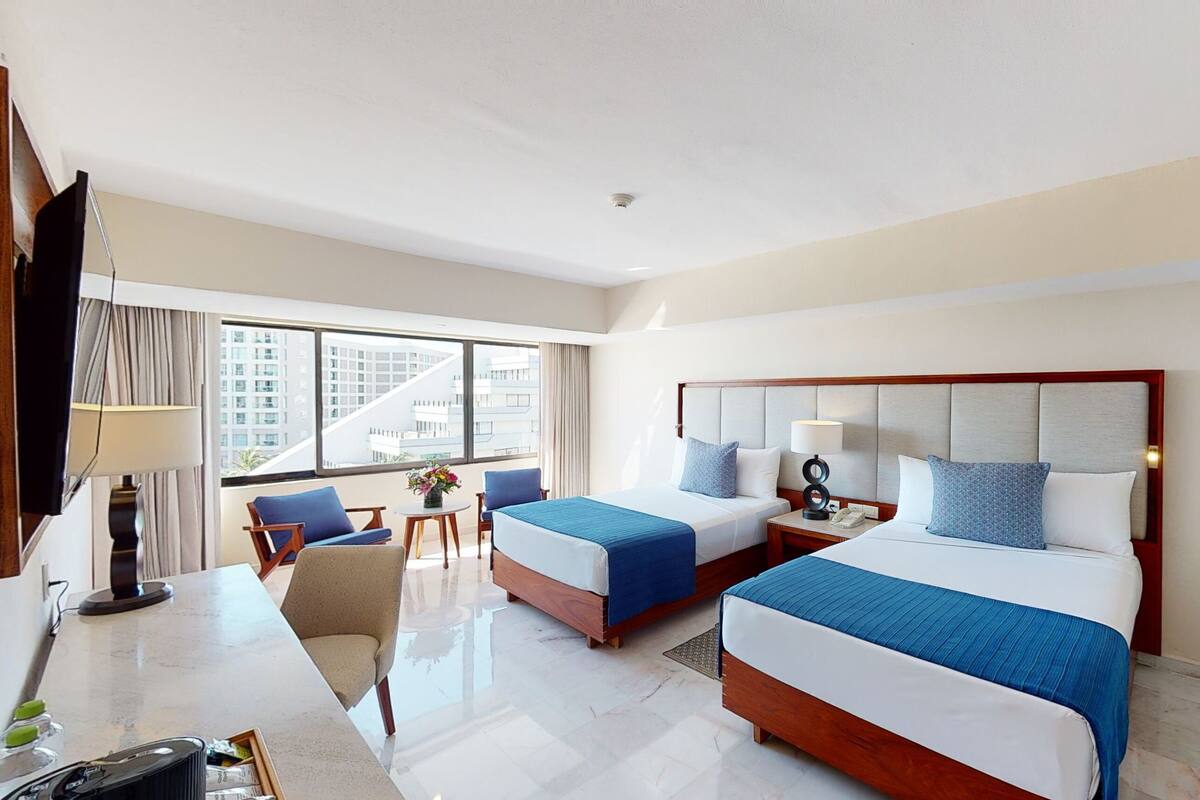 A hotel room with two beds, a desk, a chair, a TV, and a large window with a city view.