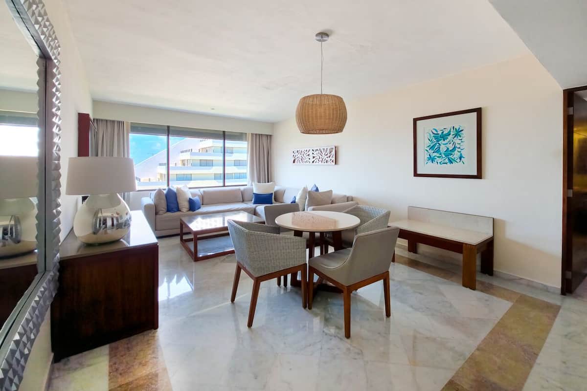 A modern hotel room with a dining area, a sofa set, a round table, and a large window offering a view of the building.