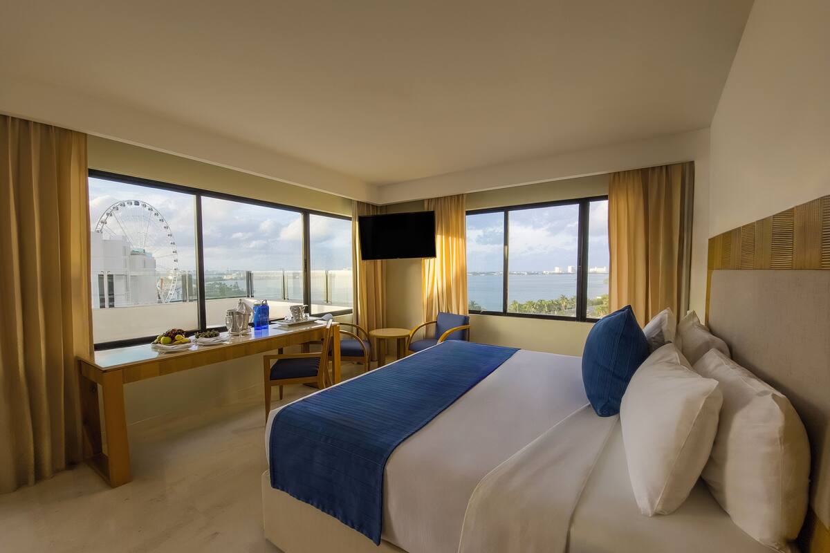 A hotel room with a bed, a desk, a chair, a TV, and large windows offering a view of a Ferris wheel and a body of water.