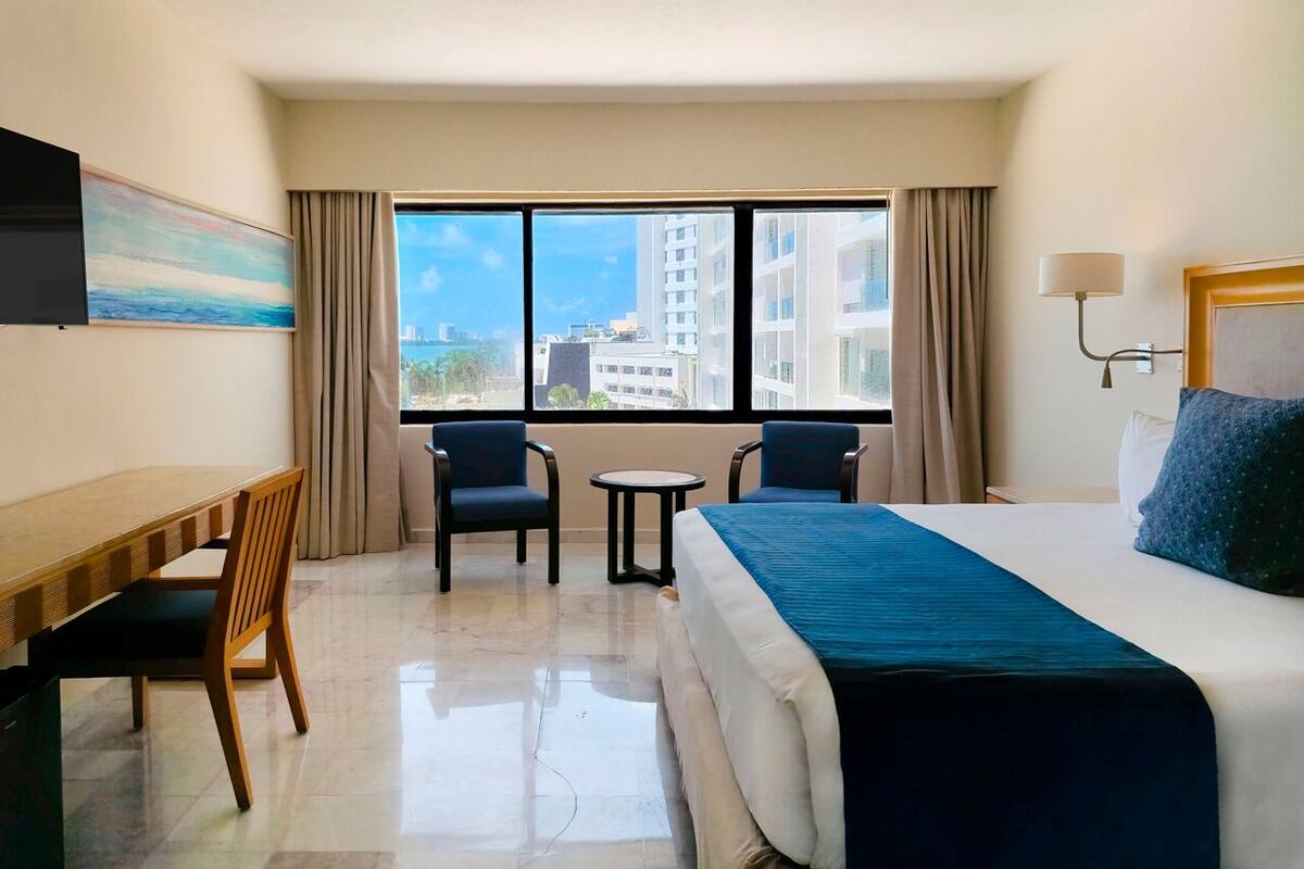 A hotel room with a bed, a desk, two chairs, a small table, and a large window offering a city view.