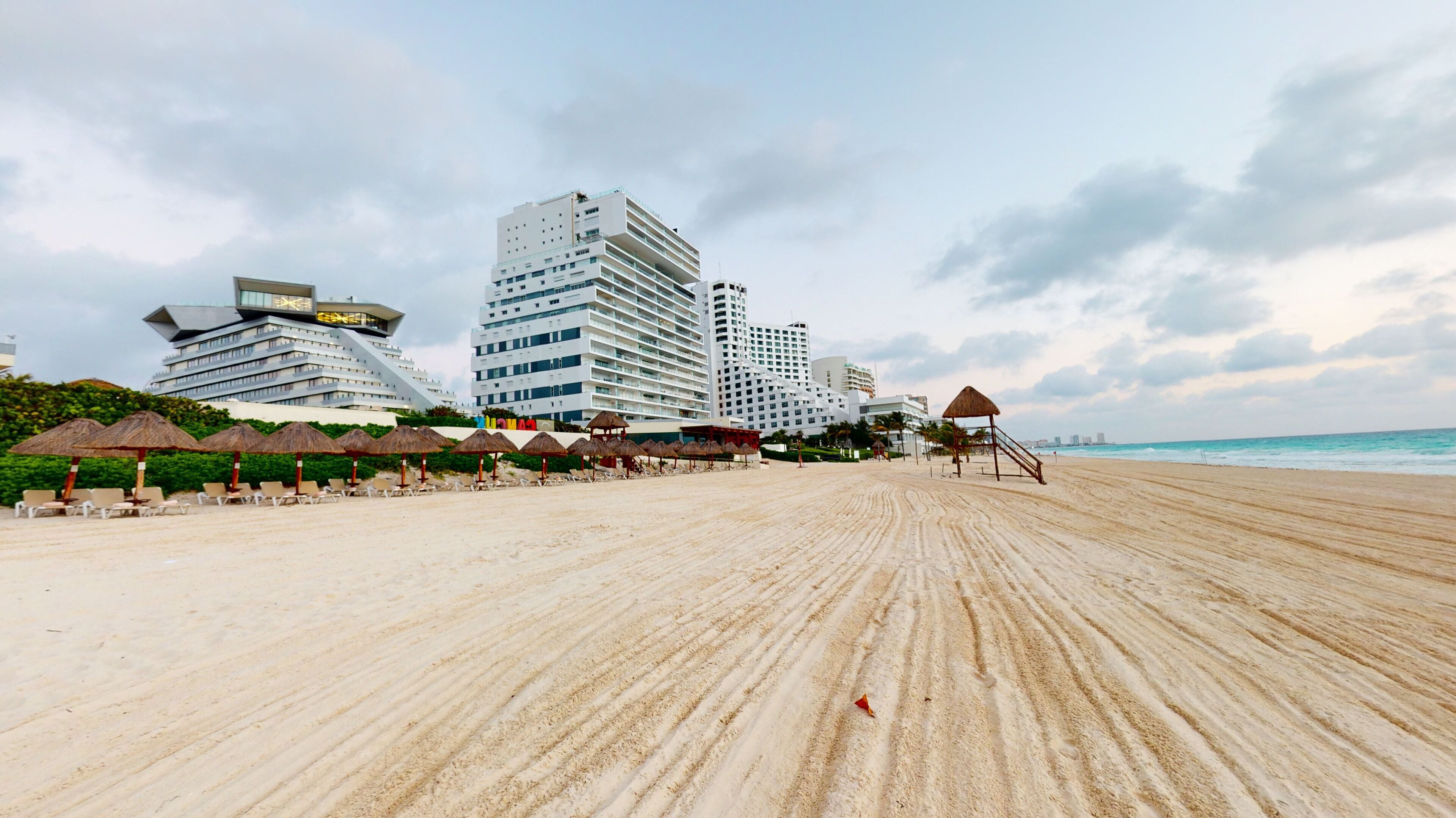 A beach with a row of sun umbrellas and lounge chairs, a thatched-roof structure, and modern high-rise buildings in the background.